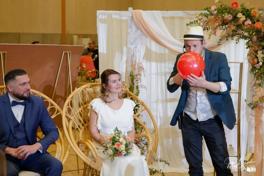 Jérôme gonfle un ballon rouge devant les mariés assis dans des fauteuils en rotin, sous un décor floral