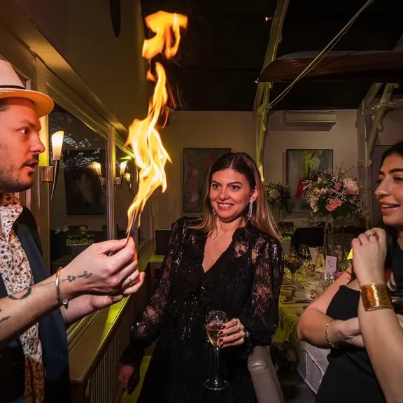 Jérôme fait jaillir une flamme d'une carte au milieu des convives, dans la salle d'un restaurant