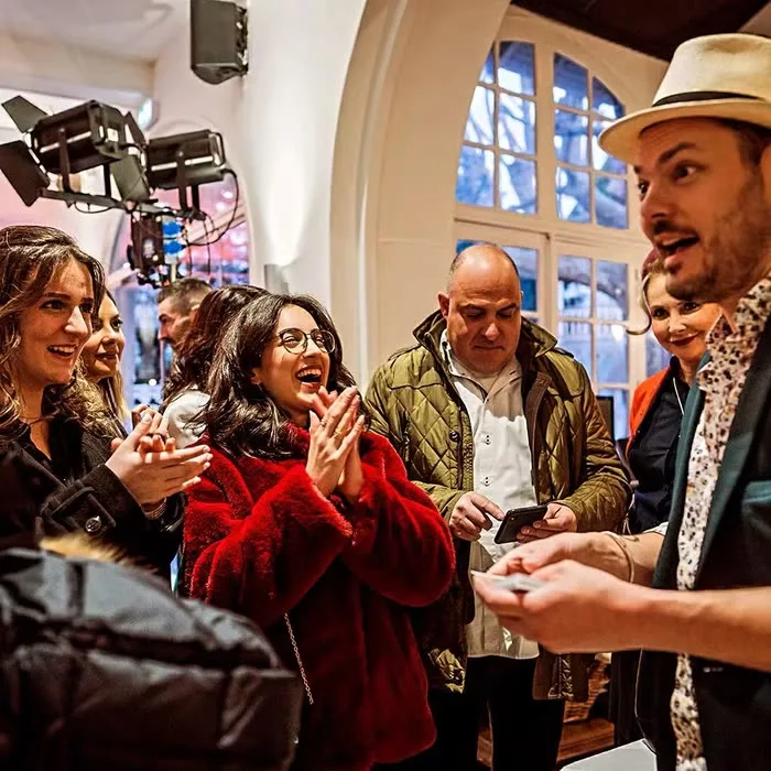 Un groupe de spectateurs éclate de rire autour de Jérôme, sous les projecteurs d’un tournage