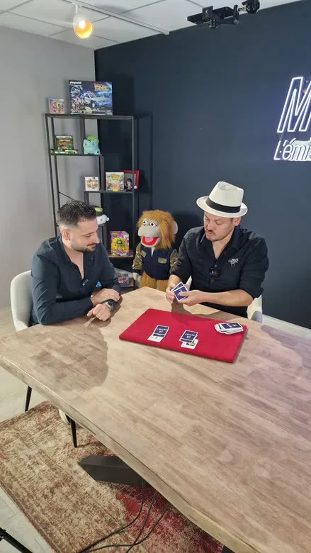Jérôme enseigne un tour de cartes à un participant, une marionnette assise entre eux
