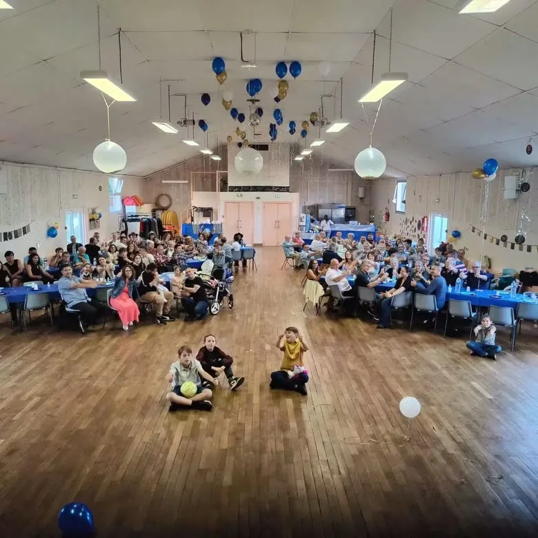Une salle des fêtes comble, enfants assis au premier rang sous des ballons bleus et dorés