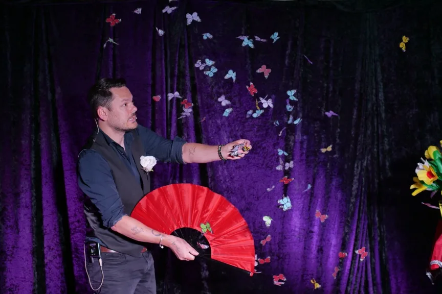 Jérôme sur scène devant un rideau de velours violet, éventail rouge en main, une nuée de papillons de couleur en suspension