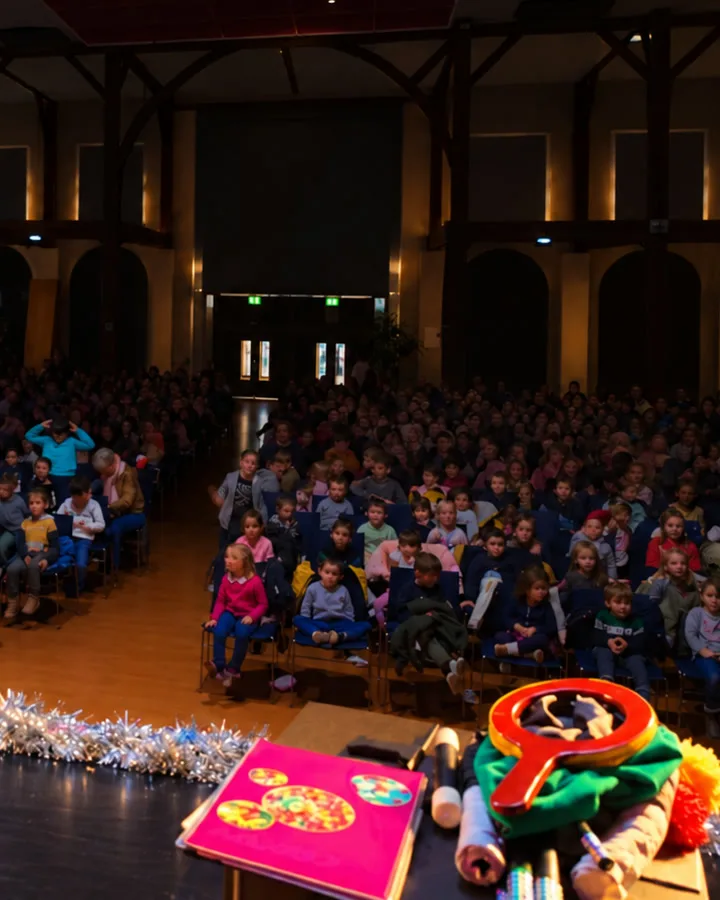 Une salle comble d’enfants vue depuis la scène, confettis et malle de magicien au premier plan
